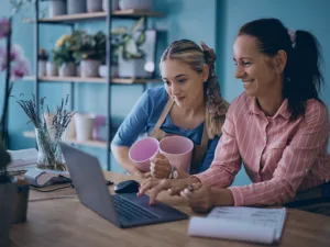Two women collaborate at a desk, looking at a laptop; one holds pink cups while the other types.