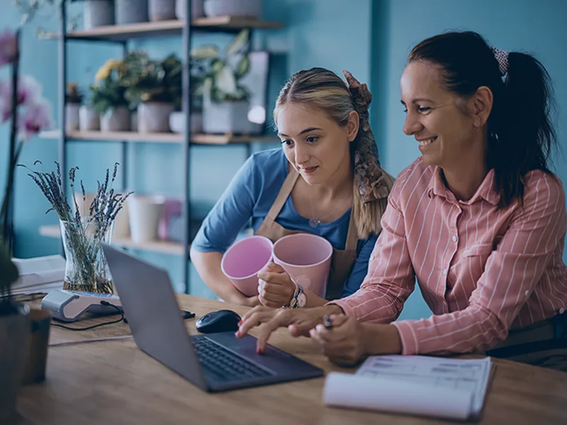 Two women collaborate at a desk, looking at a laptop; one holds pink cups while the other types.