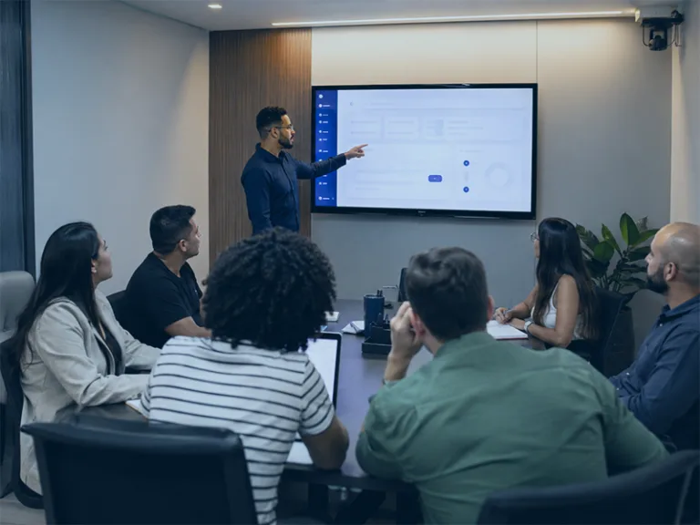 Presenter points to a large screen with a slide as six colleagues listen around a conference table in a modern boardroom.