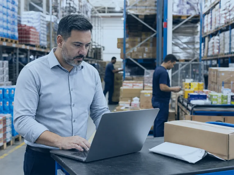 Man in a light blue shirt typing on a laptop at a warehouse packing station with boxes on shelves nearby.